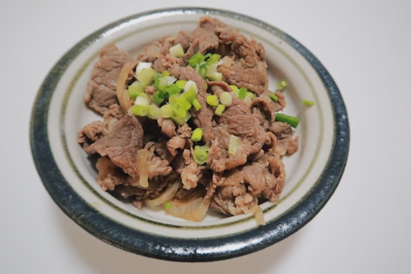 A bowl contains slices of cooked beef garnished with chopped green onions. The beef appears tender and is served with thinly sliced onions in a round ceramic dish.