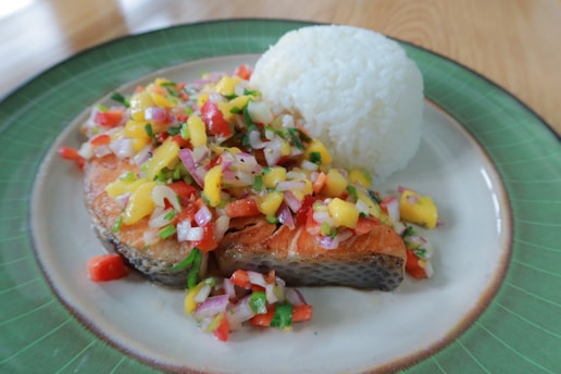 A plate of cooked fish topped with a colorful salsa made of chopped mango, red onion, tomato, and herbs, served alongside a mound of white rice. The dish is presented on a green-rimmed plate set on a table.