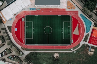 An aerial view of a large sports complex featuring a central green football field surrounded by a red running track. Adjacent to the track, there is a parking area, buildings, and pathways. Nearby, there are additional sports courts in different colors, including a blue swimming pool and orange basketball courts.