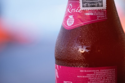 A close-up view of a chilled beverage bottle, partially obscured. The label is pink with text and a raspberry image. Condensation droplets are visible on the surface of the bottle, suggesting it is cold. The blurred background is blue and orange, giving a warm and cool contrast.