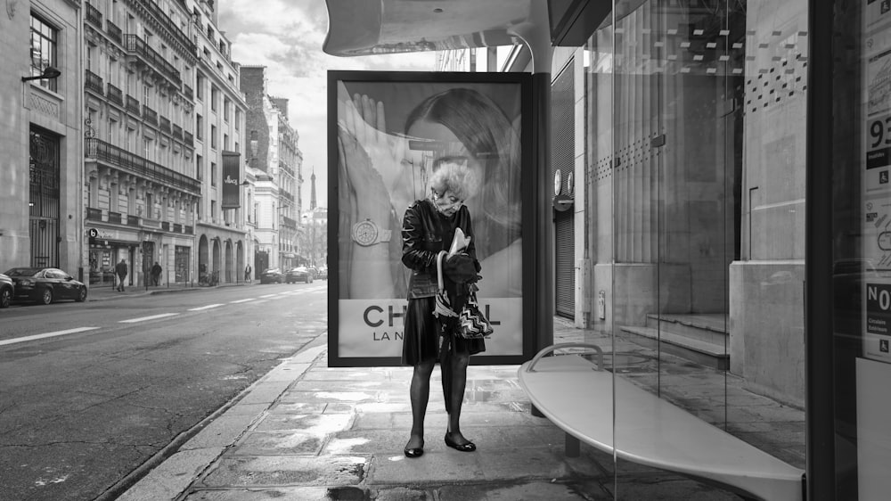 grayscale photo of woman in black coat standing near glass window