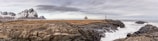 A rugged coastal landscape featuring rocky terrain and crashing ocean waves. In the distance, there are snow-capped mountains and a small weather station with a radar dome and a lighthouse, situated on the rocky land. The sky is overcast with clouds.
