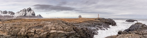 A rugged coastal landscape featuring rocky terrain and crashing ocean waves. In the distance, there are snow-capped mountains and a small weather station with a radar dome and a lighthouse, situated on the rocky land. The sky is overcast with clouds.