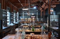 A vintage laboratory setting featuring rows of shelves filled with various glassware and scientific equipment. The room has high ceilings with exposed wooden beams and is illuminated by hanging lamps. The central workbench is cluttered with bottles, flasks, and tubes, indicating a busy and experimental atmosphere.