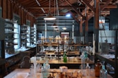 A vintage laboratory setting featuring rows of shelves filled with various glassware and scientific equipment. The room has high ceilings with exposed wooden beams and is illuminated by hanging lamps. The central workbench is cluttered with bottles, flasks, and tubes, indicating a busy and experimental atmosphere.
