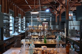 A vintage laboratory setting featuring rows of shelves filled with various glassware and scientific equipment. The room has high ceilings with exposed wooden beams and is illuminated by hanging lamps. The central workbench is cluttered with bottles, flasks, and tubes, indicating a busy and experimental atmosphere.