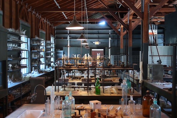 A vintage laboratory setting featuring rows of shelves filled with various glassware and scientific equipment. The room has high ceilings with exposed wooden beams and is illuminated by hanging lamps. The central workbench is cluttered with bottles, flasks, and tubes, indicating a busy and experimental atmosphere.