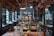 A vintage laboratory setting featuring rows of shelves filled with various glassware and scientific equipment. The room has high ceilings with exposed wooden beams and is illuminated by hanging lamps. The central workbench is cluttered with bottles, flasks, and tubes, indicating a busy and experimental atmosphere.
