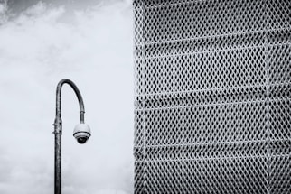 A security camera mounted on a tall, curved pole is positioned in front of a large metal mesh structure. The background features a cloudy sky, and the scene is rendered in black and white, giving it a stark, industrial feel.