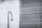 A security camera mounted on a tall, curved pole is positioned in front of a large metal mesh structure. The background features a cloudy sky, and the scene is rendered in black and white, giving it a stark, industrial feel.