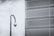 A security camera mounted on a tall, curved pole is positioned in front of a large metal mesh structure. The background features a cloudy sky, and the scene is rendered in black and white, giving it a stark, industrial feel.