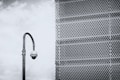 A security camera mounted on a tall, curved pole is positioned in front of a large metal mesh structure. The background features a cloudy sky, and the scene is rendered in black and white, giving it a stark, industrial feel.