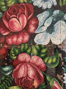 A mosaic artwork featuring large, colorful flowers. The pieces are arranged to depict rose-like flowers in shades of pink and red, white, and blue. The background contains dark and green shades representing leaves and stems.