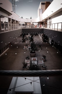 A modern outdoor shopping area with two levels, featuring a central courtyard decorated with string lights and potted plants. The upper level has railings and is flanked by stores, while the lower level has seating arrangements. A tall sign reading 'CENTURY' is prominently displayed on the right-hand side. The area appears well-maintained and designed for leisure and social activities.