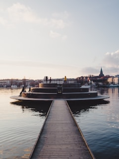 A serene waterfront scene featuring a circular platform protruding into the water, accessible via a narrow wooden walkway. Several people are seen leisurely standing or walking on the platform, with some bicycles nearby. The background consists of a calm body of water reflecting the surroundings and a cityscape with buildings and a church steeple.
