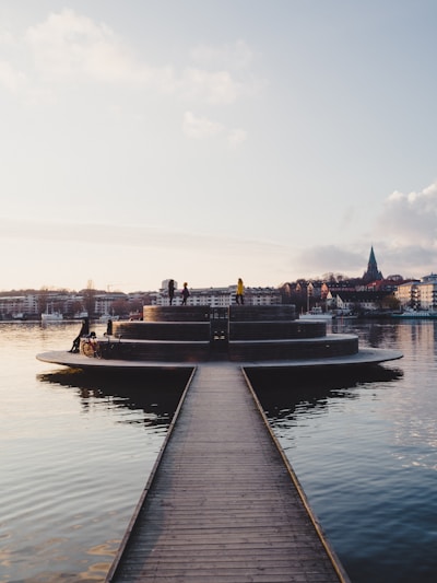 A serene waterfront scene featuring a circular platform protruding into the water, accessible via a narrow wooden walkway. Several people are seen leisurely standing or walking on the platform, with some bicycles nearby. The background consists of a calm body of water reflecting the surroundings and a cityscape with buildings and a church steeple.