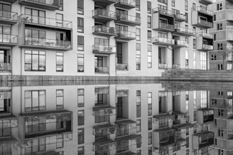A modern apartment building with numerous balconies is reflected in a calm body of water. The black and white image highlights the architectural lines and the symmetrical reflection.