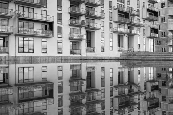 A modern apartment building with numerous balconies is reflected in a calm body of water. The black and white image highlights the architectural lines and the symmetrical reflection.
