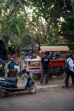 A small street food cart with an orange canopy is surrounded by people enjoying snacks. The cart has a display case with various fried items, and a stack of buns on top. Several men are sitting and standing around, some talking and others eating. A scooter is parked nearby. The setting is outdoors with lush green trees in the background creating a shaded area, suggesting a casual and lively atmosphere.