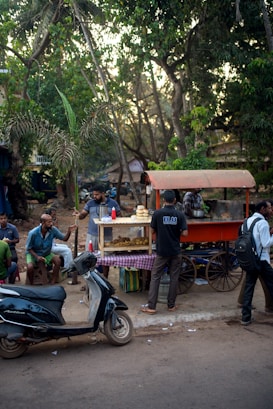 A small street food cart with an orange canopy is surrounded by people enjoying snacks. The cart has a display case with various fried items, and a stack of buns on top. Several men are sitting and standing around, some talking and others eating. A scooter is parked nearby. The setting is outdoors with lush green trees in the background creating a shaded area, suggesting a casual and lively atmosphere.