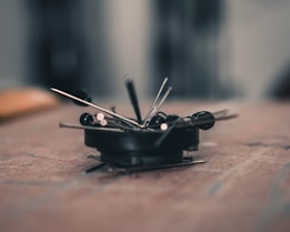 A close-up of several sewing pins and needles scattered and clustered together on a flat surface. The focus is sharp on the pins, while the background is softly blurred, creating a bokeh effect.