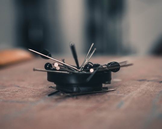 A close-up of several sewing pins and needles scattered and clustered together on a flat surface. The focus is sharp on the pins, while the background is softly blurred, creating a bokeh effect.
