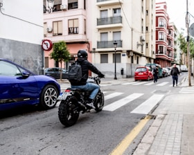 A motorcyclist wearing a helmet and backpack rides across an urban street at a crosswalk. A blue car is parked nearby, and people walk in the background. Buildings with balconies line the street, reflecting a city environment.
