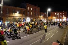 A large group of people gathered on a city street at night, many wearing bright yellow and orange reflective vests. They appear to be participating in a cycling event or protest, with numerous bicycles visible. The street is lined with historic brick buildings, and streetlights illuminate the scene.