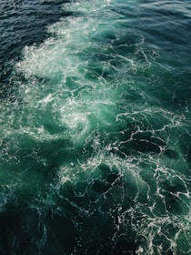 Churning ocean waves create intricate patterns of foam on a vibrant teal and deep blue sea surface. The interplay of light and water conveys a sense of movement and energy.
