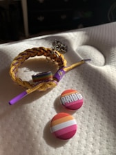 A braided bracelet with yellow and purple strands is laid on a white textured surface. Attached to the bracelet is a charm with interlocking Venus symbols. Beside the bracelet are two round buttons, one with the word 'PRIDE' written in bold white letters against a background of warm colors, and the other featuring pink, orange, and white stripes.