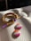 A braided bracelet with yellow and purple strands is laid on a white textured surface. Attached to the bracelet is a charm with interlocking Venus symbols. Beside the bracelet are two round buttons, one with the word 'PRIDE' written in bold white letters against a background of warm colors, and the other featuring pink, orange, and white stripes.