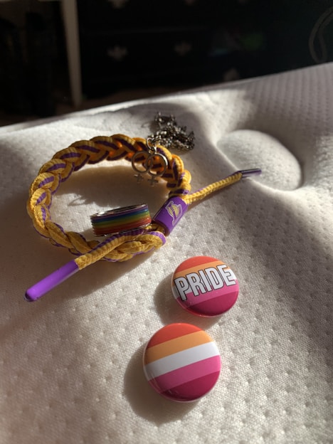 A braided bracelet with yellow and purple strands is laid on a white textured surface. Attached to the bracelet is a charm with interlocking Venus symbols. Beside the bracelet are two round buttons, one with the word 'PRIDE' written in bold white letters against a background of warm colors, and the other featuring pink, orange, and white stripes.