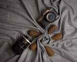 A vintage-style camera and a camera lens are artfully arranged on a textured gray fabric with three decorative pine cones placed around them. The fabric is gathered at the center to create a visually pleasing display.