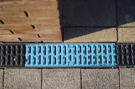 A close-up view of a street drainage system featuring a blue metal grate with embossed text, surrounded by pavement and a brick wall. The grate design includes elongated slots for water drainage.