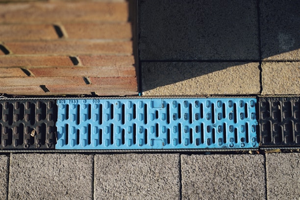 A close-up view of a street drainage system featuring a blue metal grate with embossed text, surrounded by pavement and a brick wall. The grate design includes elongated slots for water drainage.