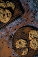 A dark, moody setting showcases a variety of chocolate chip cookies on two plates. Cocoa powder and chocolate chips are scattered artistically on a dark, textured surface. One of the cookies is partially broken, adding a casual touch to the arrangement.