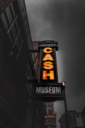 A vintage-style neon sign prominently features the word 'CASH' in bold, illuminated letters against a moody, overcast sky. The sign is part of a larger advertisement for a museum, likely a music or cultural landmark. The surrounding buildings are dark, creating a stark contrast with the bright sign, adding an urban atmospheric touch.