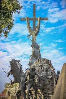 A large monument depicting a group of sculpted figures in dynamic poses, with a prominent cross and stylized sails behind them. The sculpture is set against a vibrant blue sky with scattered clouds and framed by leafy green branches. The figures appear to be in motion, characterized by dramatic expressions and flowing garments.