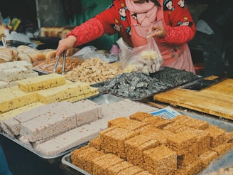 A vendor is arranging a variety of colorful snacks or sweets on a market table. Various types of confections, including blocks of sesame, rice, and nut-based treats, are neatly displayed in trays. The vendor is wearing a red jacket with cartoon patterns and a scarf, and is using tongs to place an item into a plastic bag.