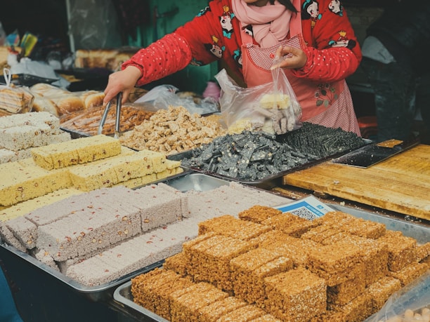 A vendor is arranging a variety of colorful snacks or sweets on a market table. Various types of confections, including blocks of sesame, rice, and nut-based treats, are neatly displayed in trays. The vendor is wearing a red jacket with cartoon patterns and a scarf, and is using tongs to place an item into a plastic bag.