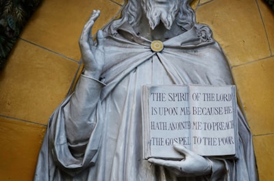 A stone statue features a bearded figure holding a book with engraved text. The text reads: 'THE SPIRIT OF THE LORD IS UPON ME BECAUSE HE HATH ANOINTED ME TO PREACH THE GOSPEL TO THE POOR.' The figure's hand is raised in a gesture, and the statue is set against a mustard-colored background.