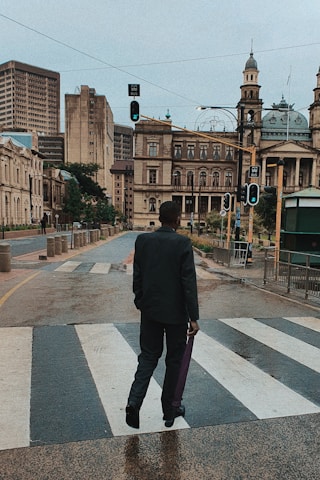 A person dressed in a suit walks across a wet pedestrian crossing in an urban area, with historic buildings and modern skyscrapers in the background. The streetlights are green, indicating it's safe to cross.