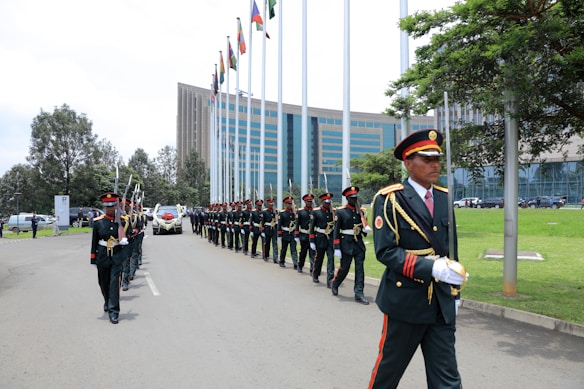 A formal procession involves a group of uniformed personnel marching in formation next to a line of tall flagpoles displaying various national flags. In the foreground, a senior member in distinct attire with gold accents leads the formation. A large modern building with glass windows serves as the backdrop, with neatly manicured green lawns and trees lining the road.