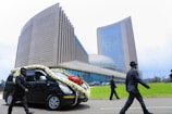 A hearse adorned with white and red flowers drives past a large, modern building with tall glass windows and a distinctive architectural design. The scene includes several people dressed formally in black suits and hats walking nearby.
