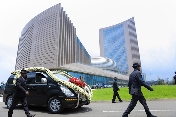 A hearse adorned with white and red flowers drives past a large, modern building with tall glass windows and a distinctive architectural design. The scene includes several people dressed formally in black suits and hats walking nearby.