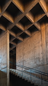 A concrete staircase runs at an angle alongside a large, textured concrete wall. The wall features a geometric pattern of recessed squares, creating a grid-like appearance across the ceiling area. The structure is characterized by its modern, industrial look.