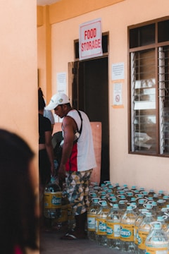 Several people are in a room labeled 'Food Storage', with large bottles of liquid stacked on the floor. One person is picking up a bottle.