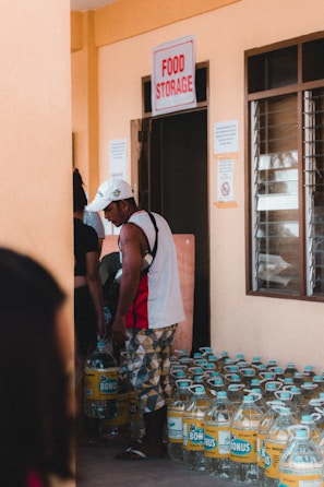 Several people are in a room labeled 'Food Storage', with large bottles of liquid stacked on the floor. One person is picking up a bottle.