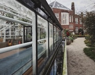 A red brick building with large windows stands adjacent to a glass greenhouse structure. The greenhouse features white metal piping and clear glass panels. A pathway lined with greenery and planters leads towards the building, with a few people visible in the distance. The sky is overcast, adding a moody ambiance.