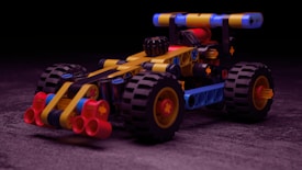 A colorful toy car built from interlocking plastic bricks, featuring large black wheels with red accents. The body of the car includes elements in yellow, red, black, and blue, and it is set against a dark, subtly textured background.